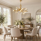 The Clérachic chandelier lit above a round table with cream upholstered chairs, in a bright room with tall windows and an olive tree.