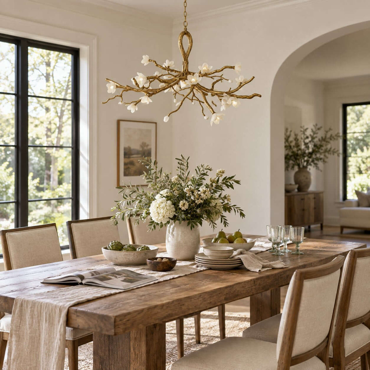 Close view of the gold branches and ceramic blossoms on this Clérachic chandelier, above a rustic table laid with a runner and a magazine.