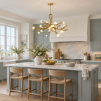 A Clérachic gold chandelier lit above a blue-grey kitchen island with woven stools, beside a window and marble splashback.