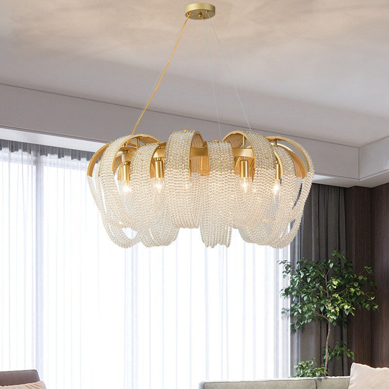 Clérachic round gold chandelier with draped crystal bead loops, hung in a bright living room beside sheer curtains and a potted tree.