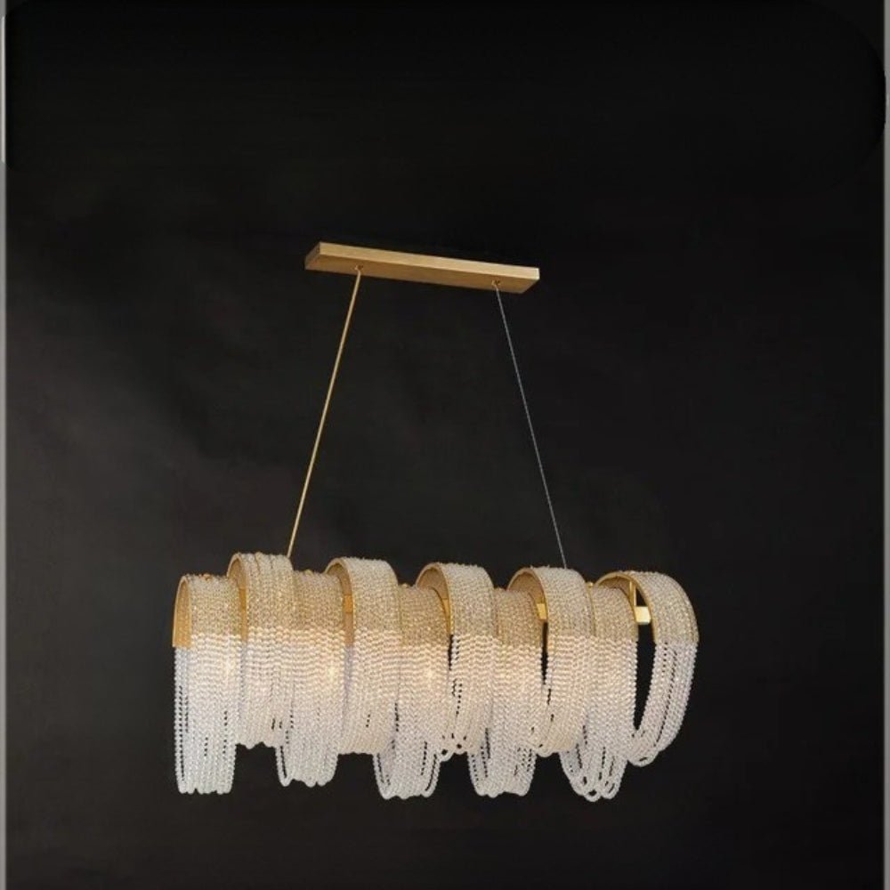 Clérachic linear gold chandelier with looping crystal bead chains hung from a rectangular brass canopy on a dark background.