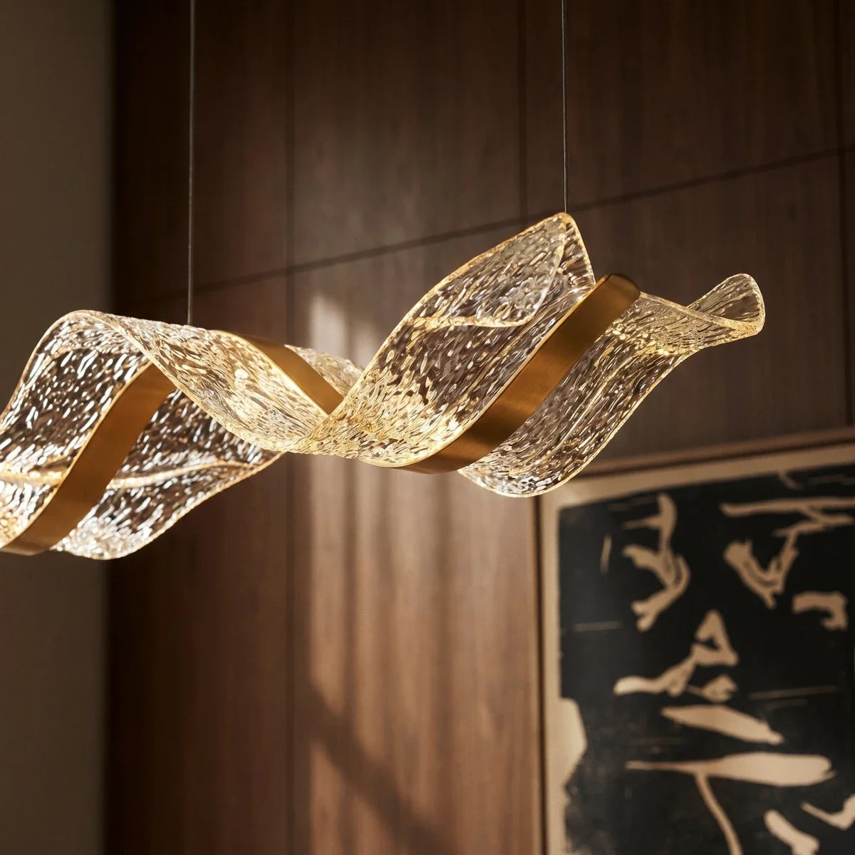 Close-up of the lit Clérachic chandelier's gold ribbon form against dark walnut wall panelling.