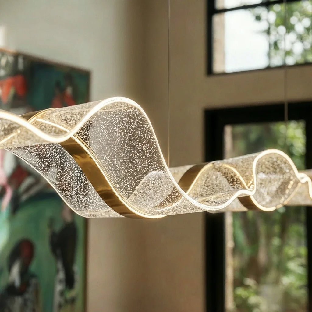 Close-up of the lit Clérachic chandelier's textured ribbon form against a leafy window backdrop.