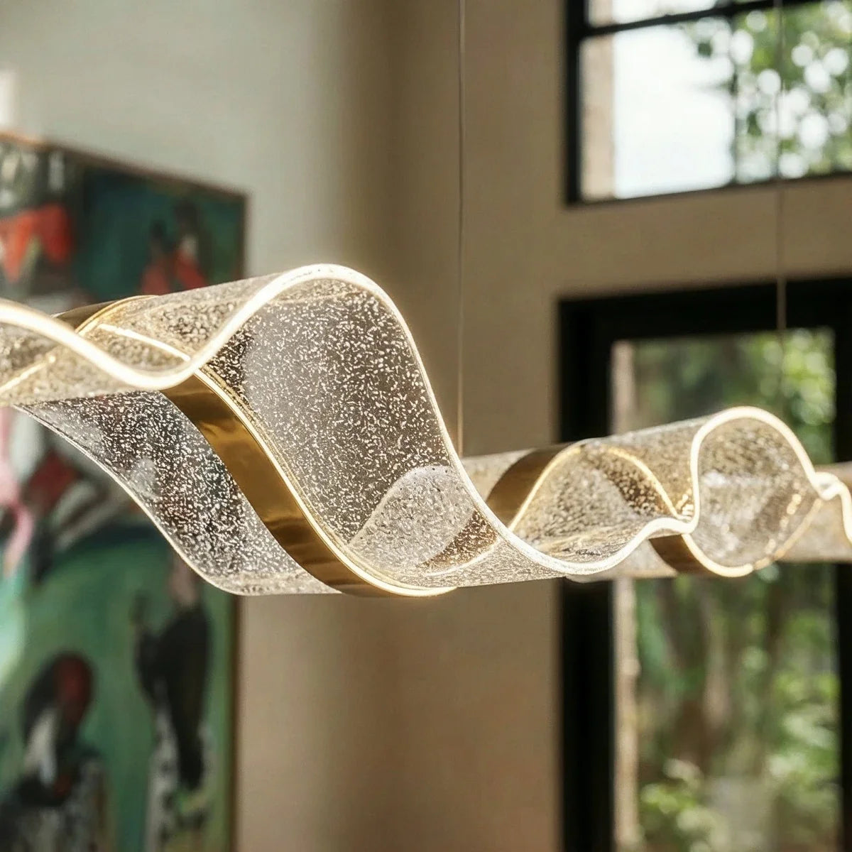 Close-up of the lit Clérachic chandelier's textured ribbon form against a leafy window backdrop.