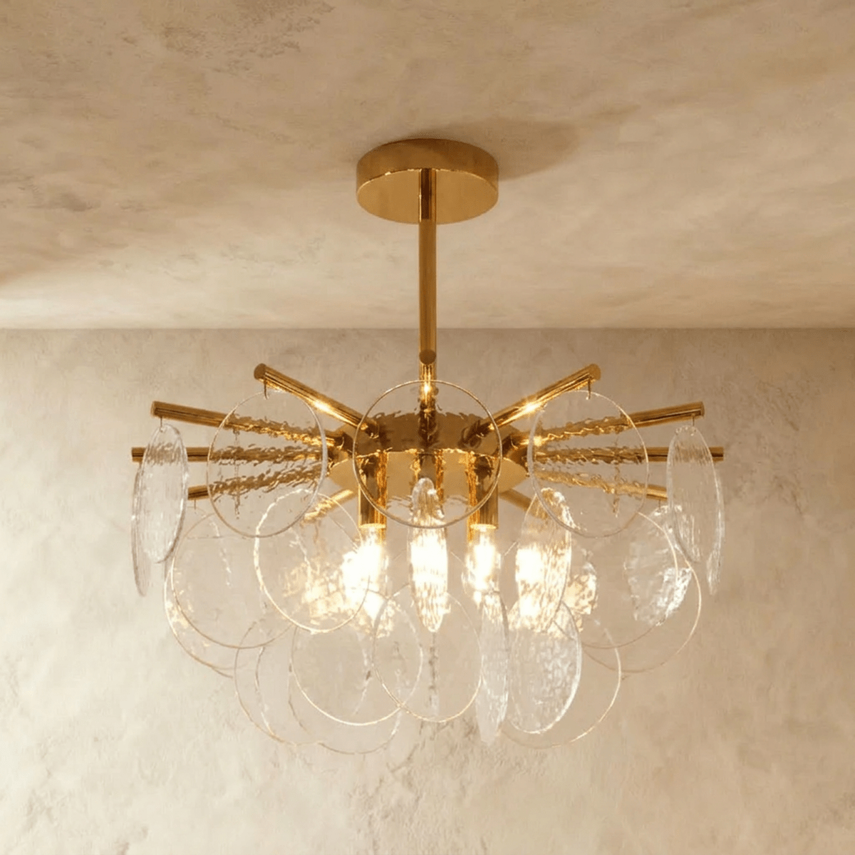 Clérachic Isla chandelier lit warm, clear transparent glass discs on brass arms, three bulbs glowing at its centre.