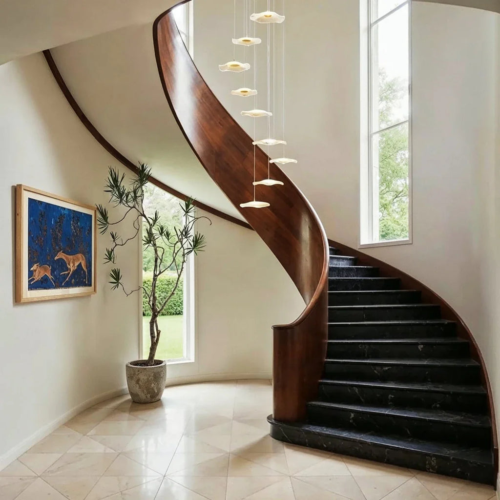 Clérachic staircase chandelier in gold cascading beside a curved wood staircase, its discs glowing above a marble floor.