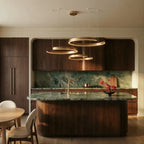 A warm gold Clérachic three-ring chandelier glows above a green marble island in a walnut kitchen.
