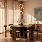 Clérachic staggered acrylic rings glowing above a round walnut dining table with olive velvet chairs and linen curtains.