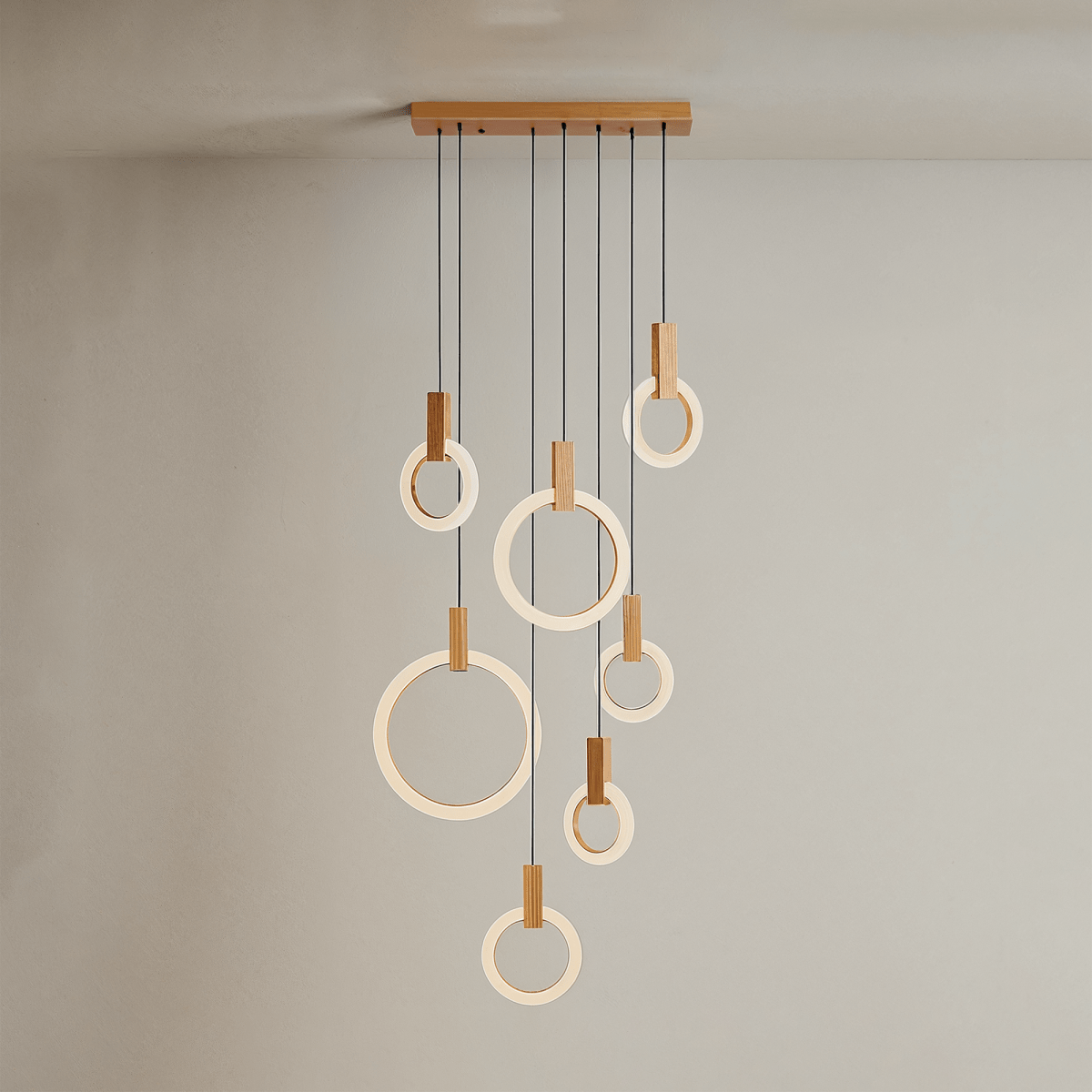 Clérachic seven lit acrylic rings with oak blocks hung from a long oak canopy on black cords against a grey wall.