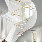 Clérachic gold six-ring LED chandelier spiralling down a curved white stairwell beside tall panelled walls and a window.