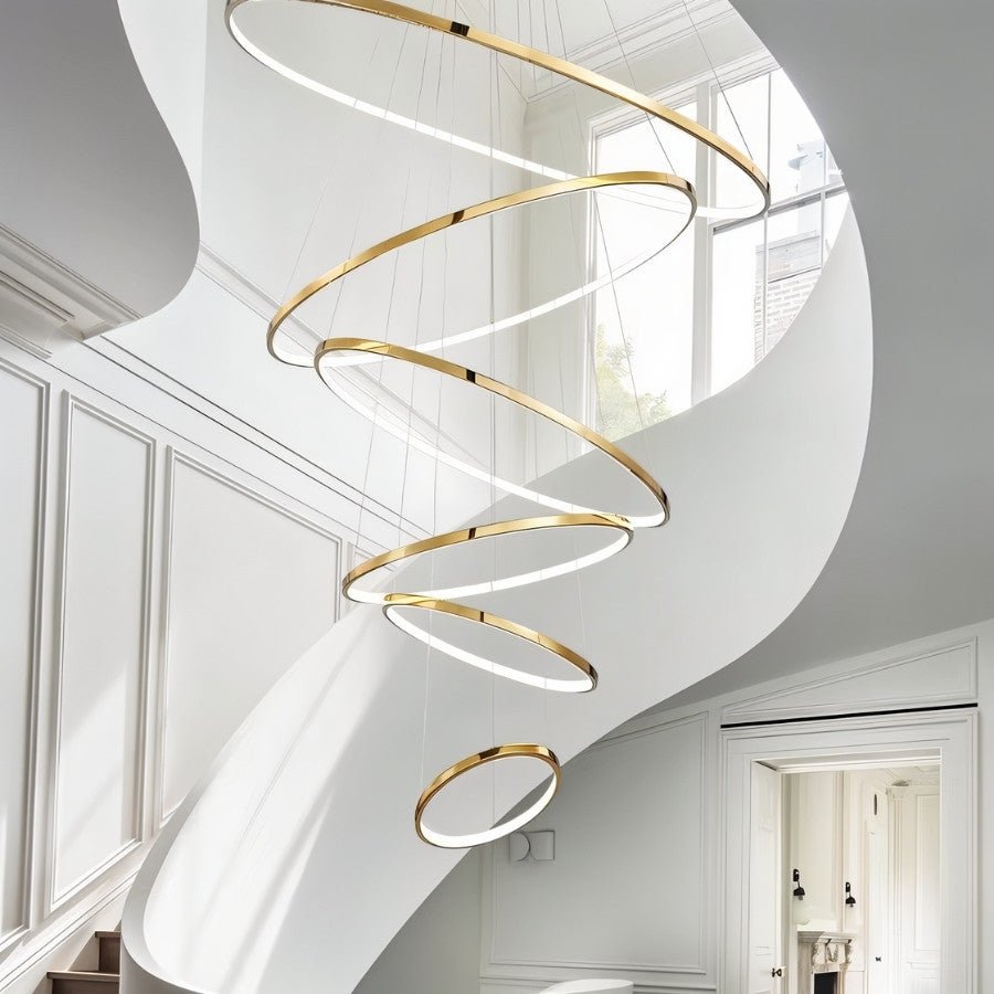 Clérachic gold six-ring LED chandelier spiralling down a curved white stairwell beside tall panelled walls and a window.