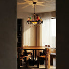 Clérachic parrot chandelier glowing over a round wooden dining table, seen through a doorway with sheer curtains beyond.