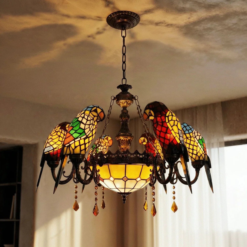 Clérachic chandelier of glass parrots with an amber bowl and crystal drops, lit warmly against a ceiling and sheer curtains.