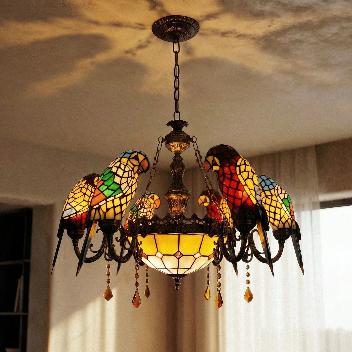 Clérachic chandelier of glass parrots with an amber bowl and crystal drops, lit warmly against a ceiling and sheer curtains.