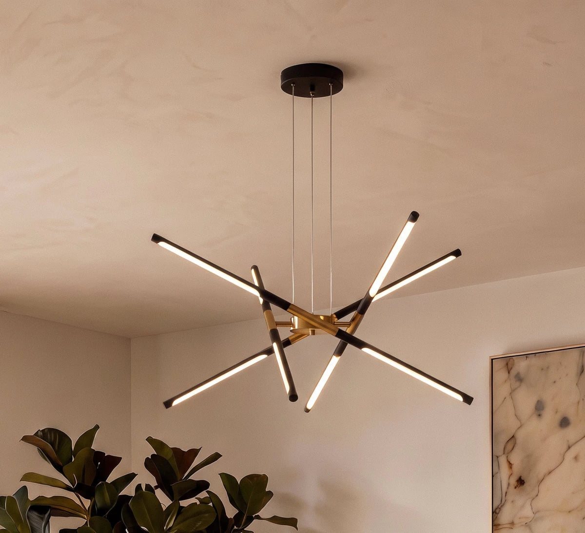 Clérachic black and gold LED rod chandelier glowing warm beneath a plaster-finish ceiling, with a leafy plant below.
