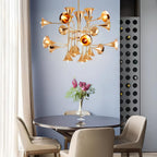 A Clérachic chandelier with gold horn-shaped shades hangs above a round dark table with a vase of pink flowers, against a blue wall.