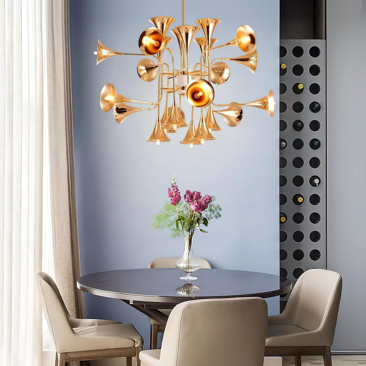 A Clérachic chandelier with gold horn-shaped shades hangs above a round dark table with a vase of pink flowers, against a blue wall.