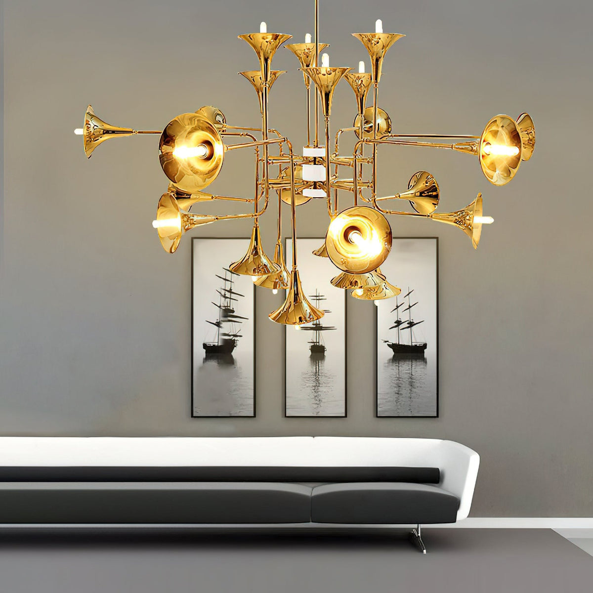 Gold trumpet-head Clérachic chandelier hung above a low grey sofa, with a triptych of ship prints on the wall behind.