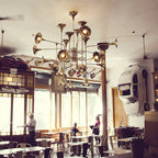 Gold Clérachic chandelier hung in a cafe with big windows, wall decor and round tables, with people at the tables below.