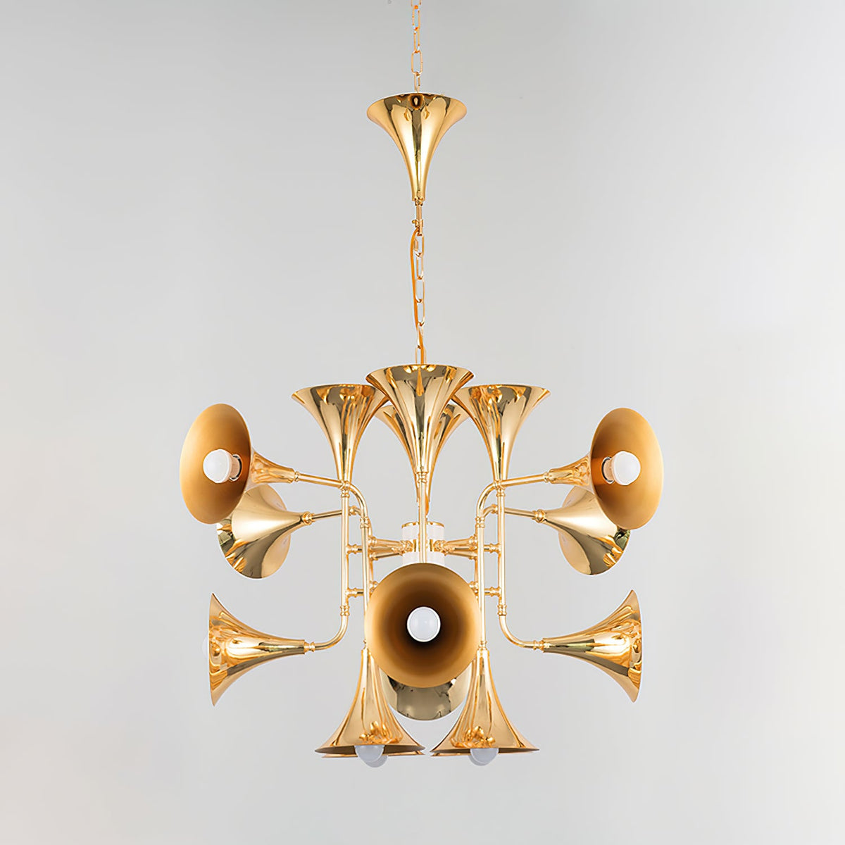 Gold trumpet-head Clérachic chandelier hanging from a chain and ceiling horn, photographed on a pale grey backdrop.