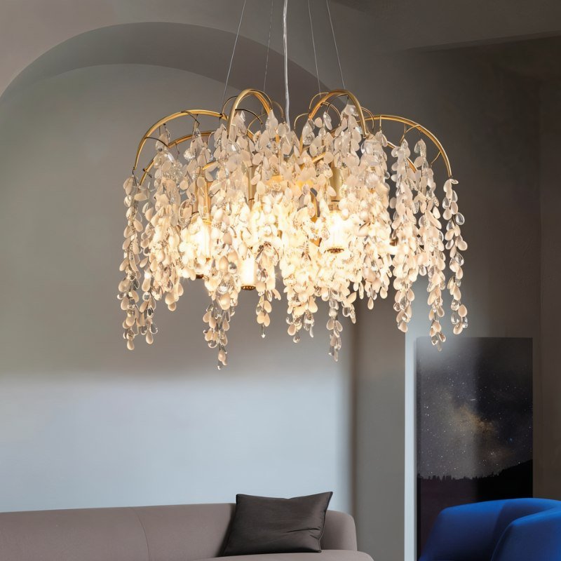 Clérachic gold crystal chandelier glowing over a sofa in a grey room, its drops catching the warm light from six bulbs