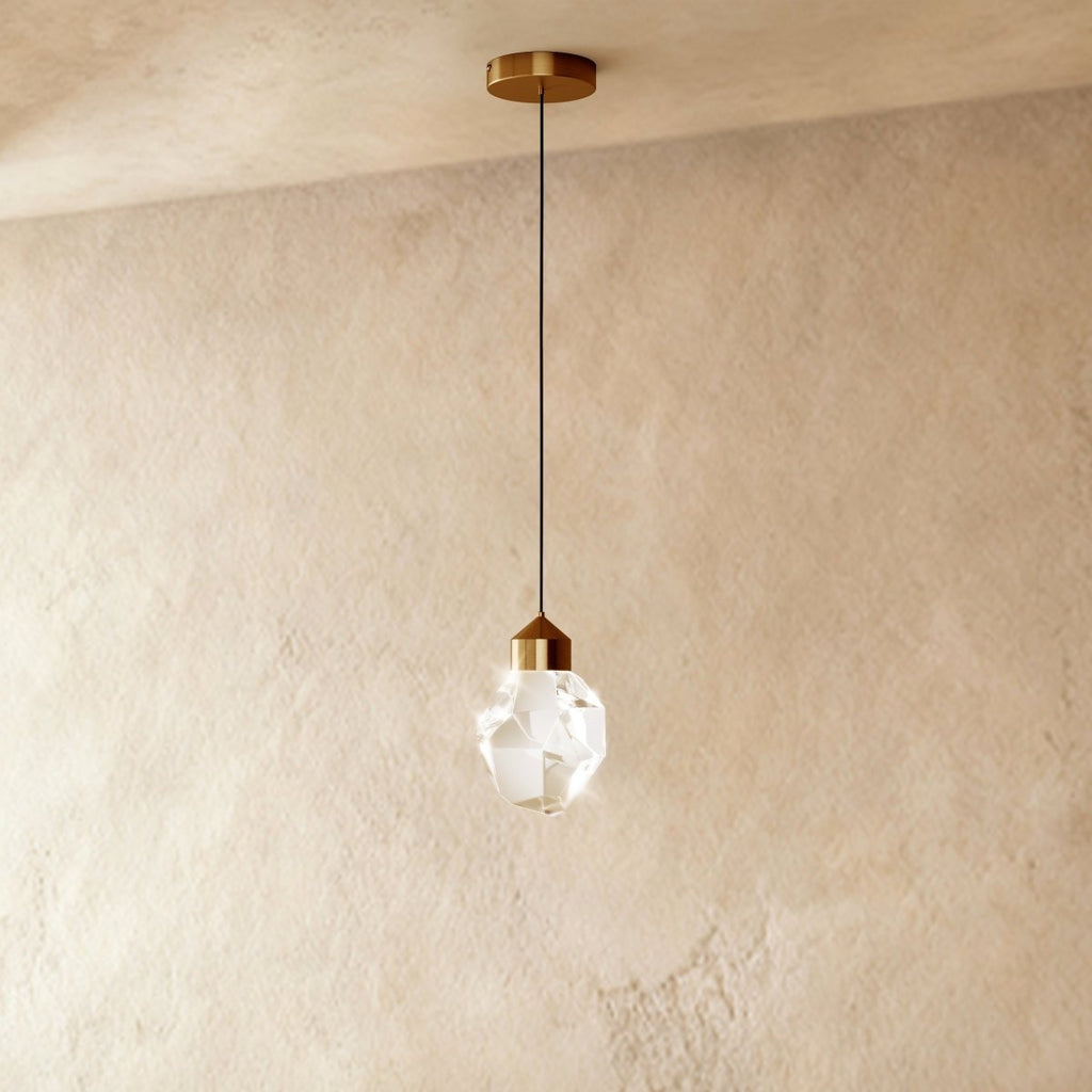 The gold crystal drop pendant light from Clérachic hangs on a brass cord straight down against a soft plaster wall.