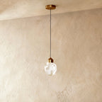 The gold crystal drop pendant light from Clérachic hangs on a brass cord straight down against a soft plaster wall.