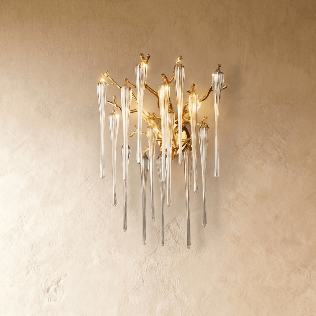Clérachic wall lamp of gold branches hung with clear glass icicle drops, lit and mounted on a textured plaster wall.