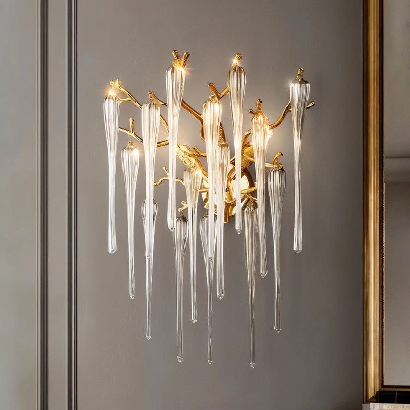 Clérachic gold branch wall lamp with glass icicle drops glowing on a grey panelled wall beside a gold-framed mirror in a room.