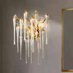 Clérachic wall lamp of gold twigs and glass icicle drops lighting a grey wall next to a slim gold-framed mirror in a room.