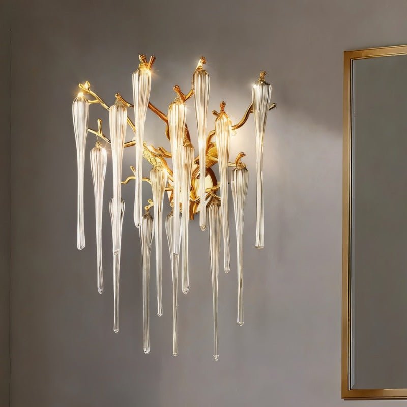 Clérachic wall lamp of gold twigs and glass icicle drops lighting a grey wall next to a slim gold-framed mirror in a room.