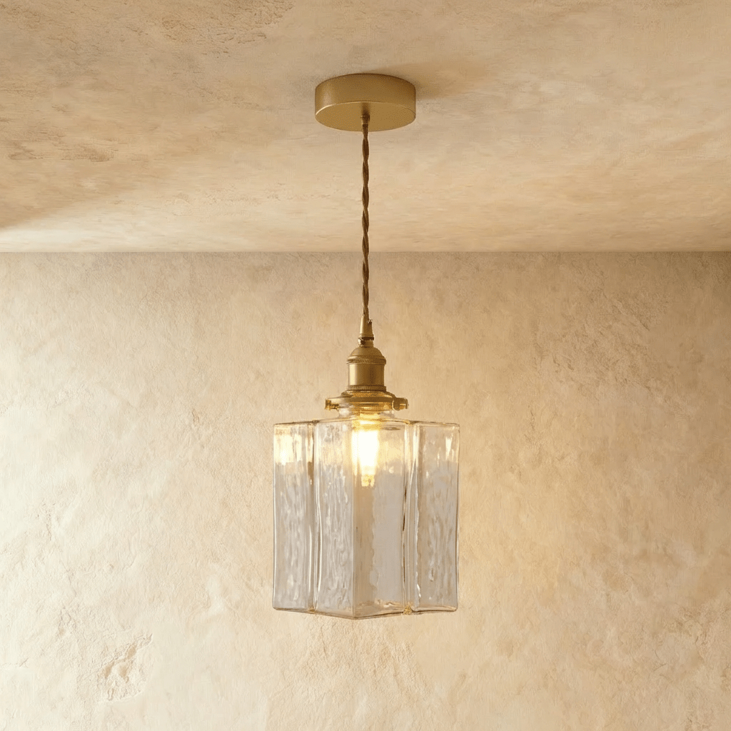 Clérachic gold cube pendant light hangs low from a twisted cord, textured glass shade lit on a plaster wall.