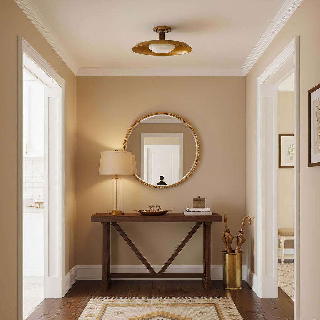 A Clérachic gold dish ceiling light above a narrow entry with a round mirror, a wooden console and a table lamp.