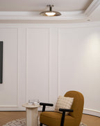 A Clérachic nickel dish ceiling light in a white panelled room with a coved ceiling, above a mustard armchair.