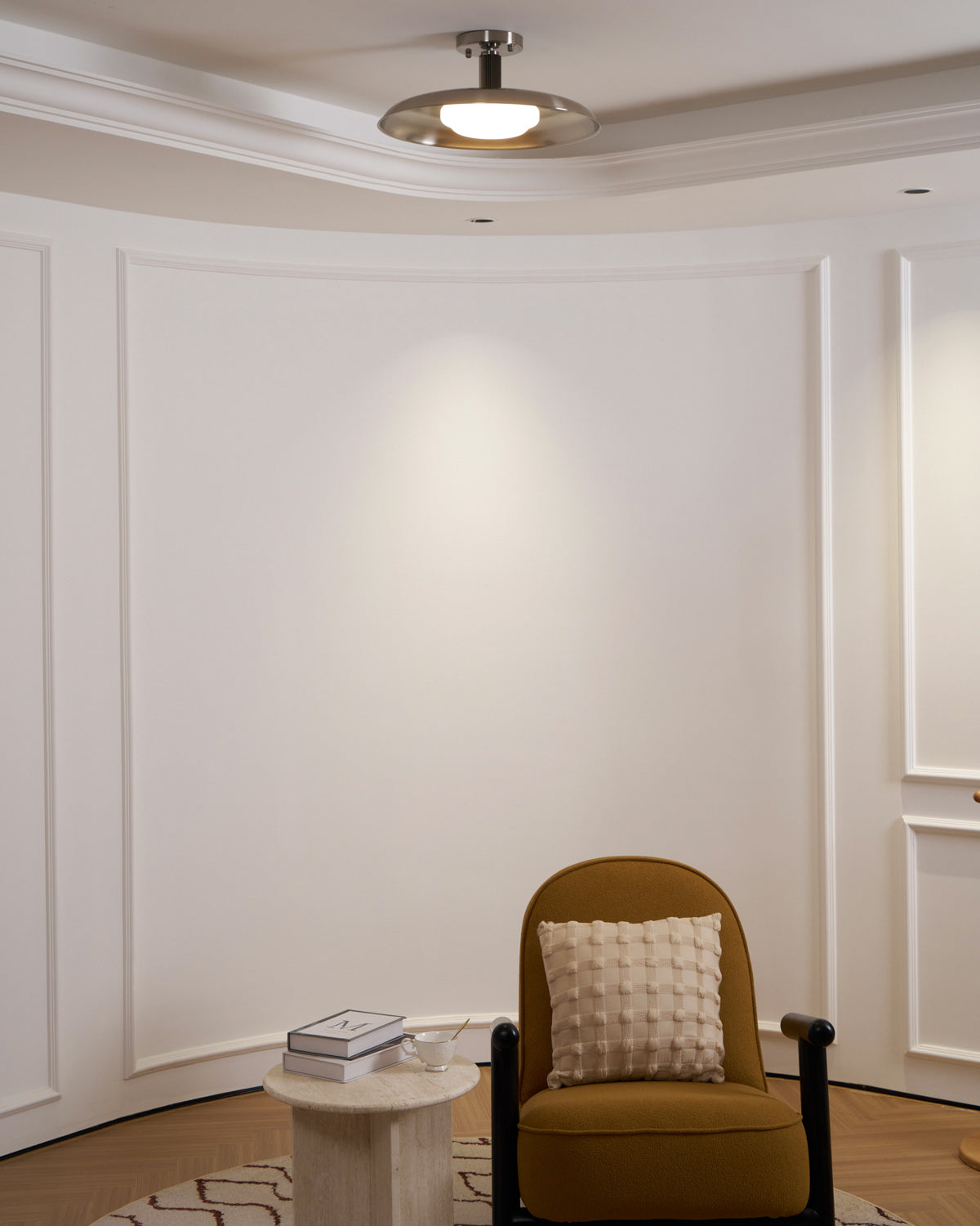 Clérachic nickel ceiling light seen from across a curved white panelled room, with a mustard chair and side table below.