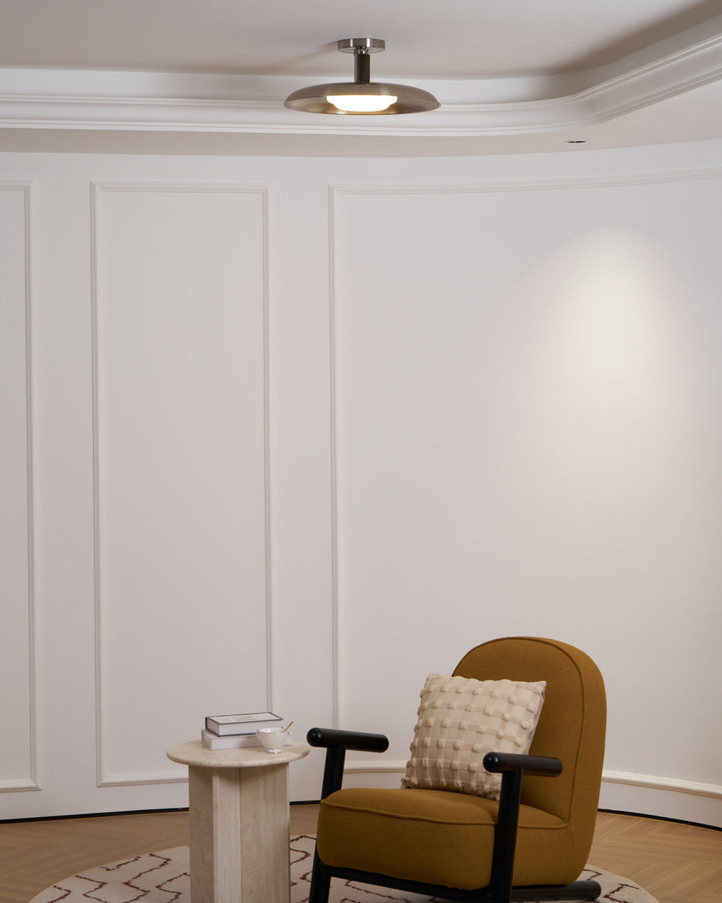 A Clérachic nickel dish ceiling light above a white panelled room, with a mustard armchair and a small stone table.