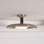 Clérachic nickel dish ceiling light with its diffuser glowing along the lower edge, seen against a white ceiling.