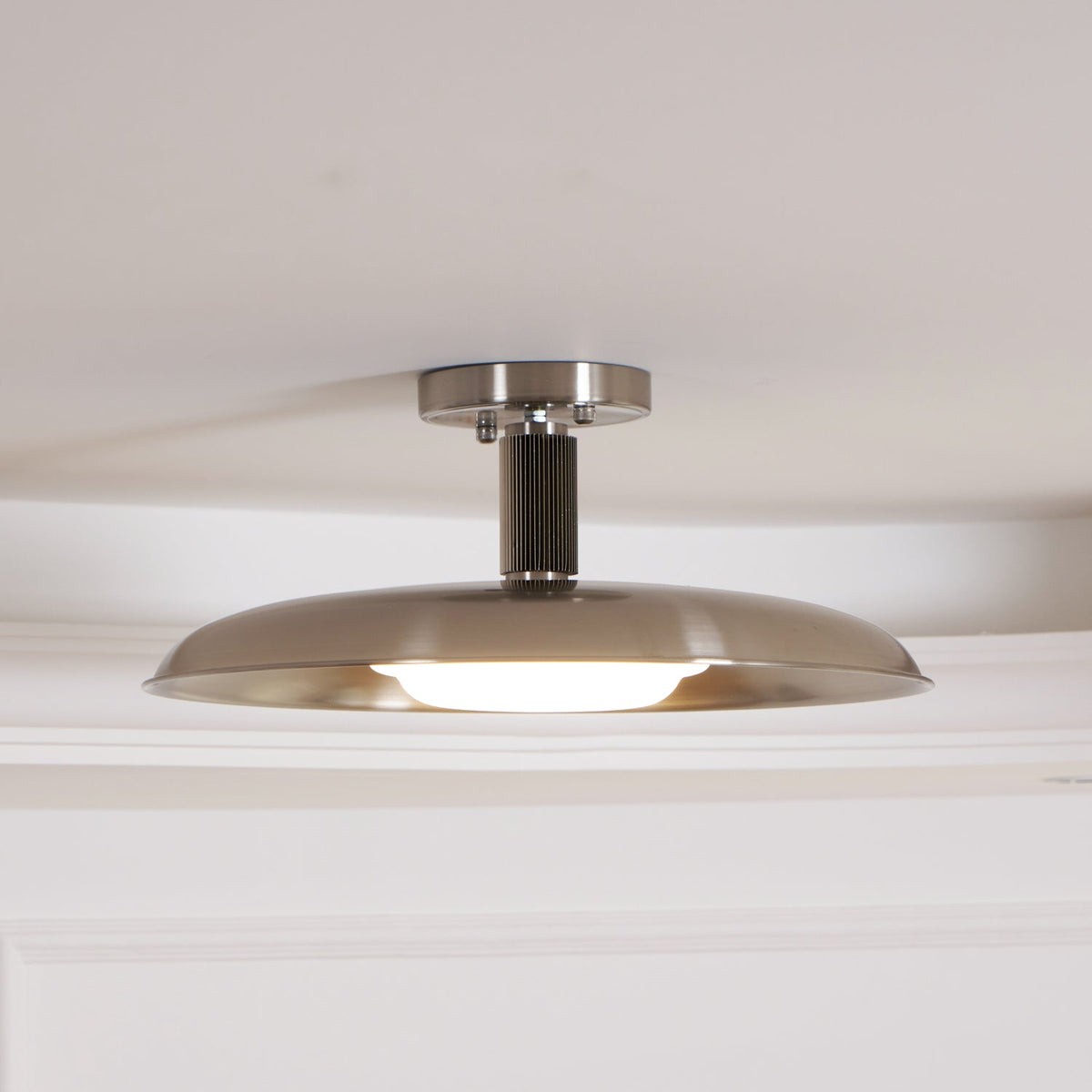 Clérachic nickel dish ceiling light with its diffuser glowing along the lower edge, seen against a white ceiling.