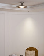 Small nickel Clérachic ceiling light seen from across a white panelled room, above a mustard chair.