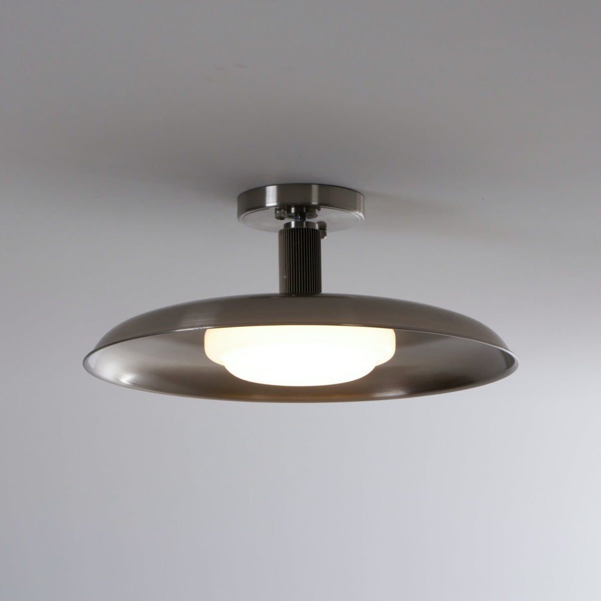 A Clérachic nickel dish ceiling light with its white diffuser lit, against a soft grey ceiling.