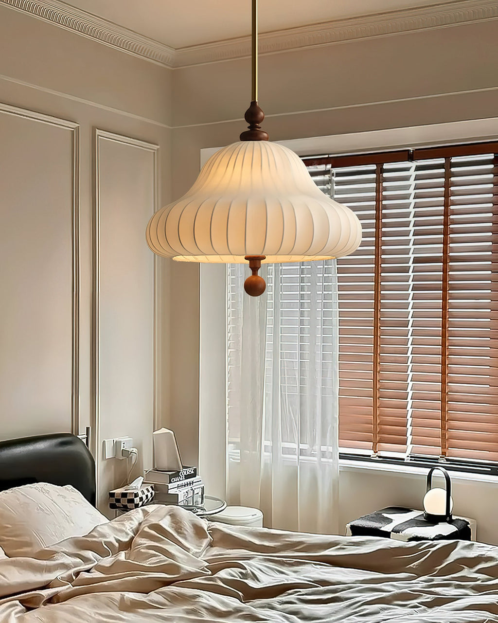 A Clérachic pendant lit in a bedroom with a panelled wall, wooden blinds, white curtains and a rumpled bed below.