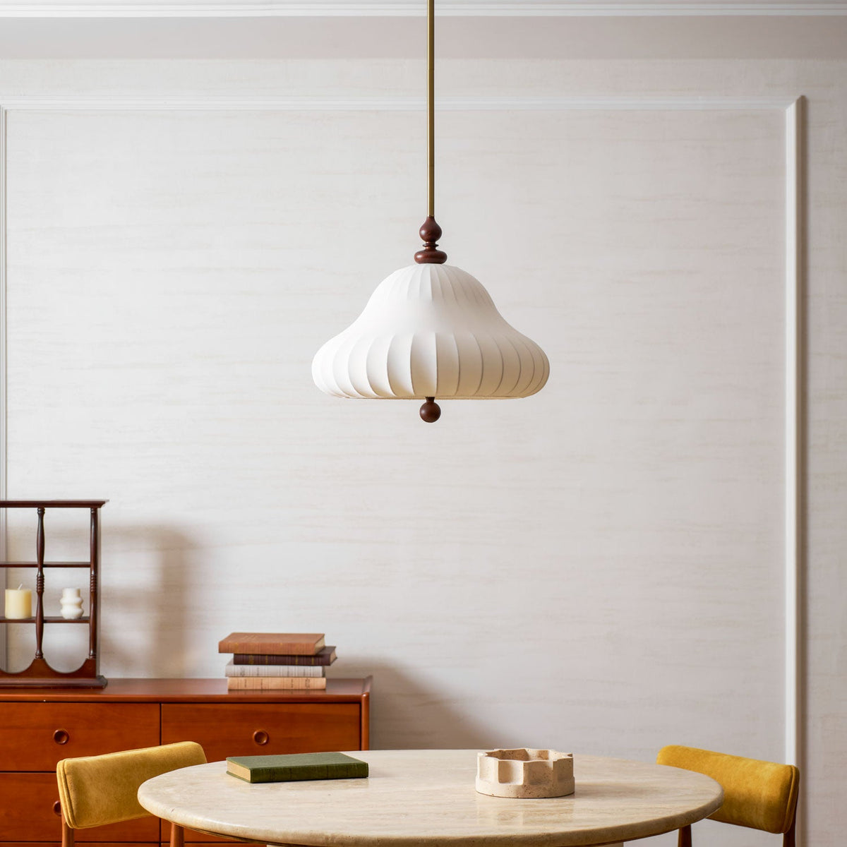 Unlit white pleated shade of this Clérachic pendant hanging against a panelled wall, with a red sideboard and a round stone table behind.