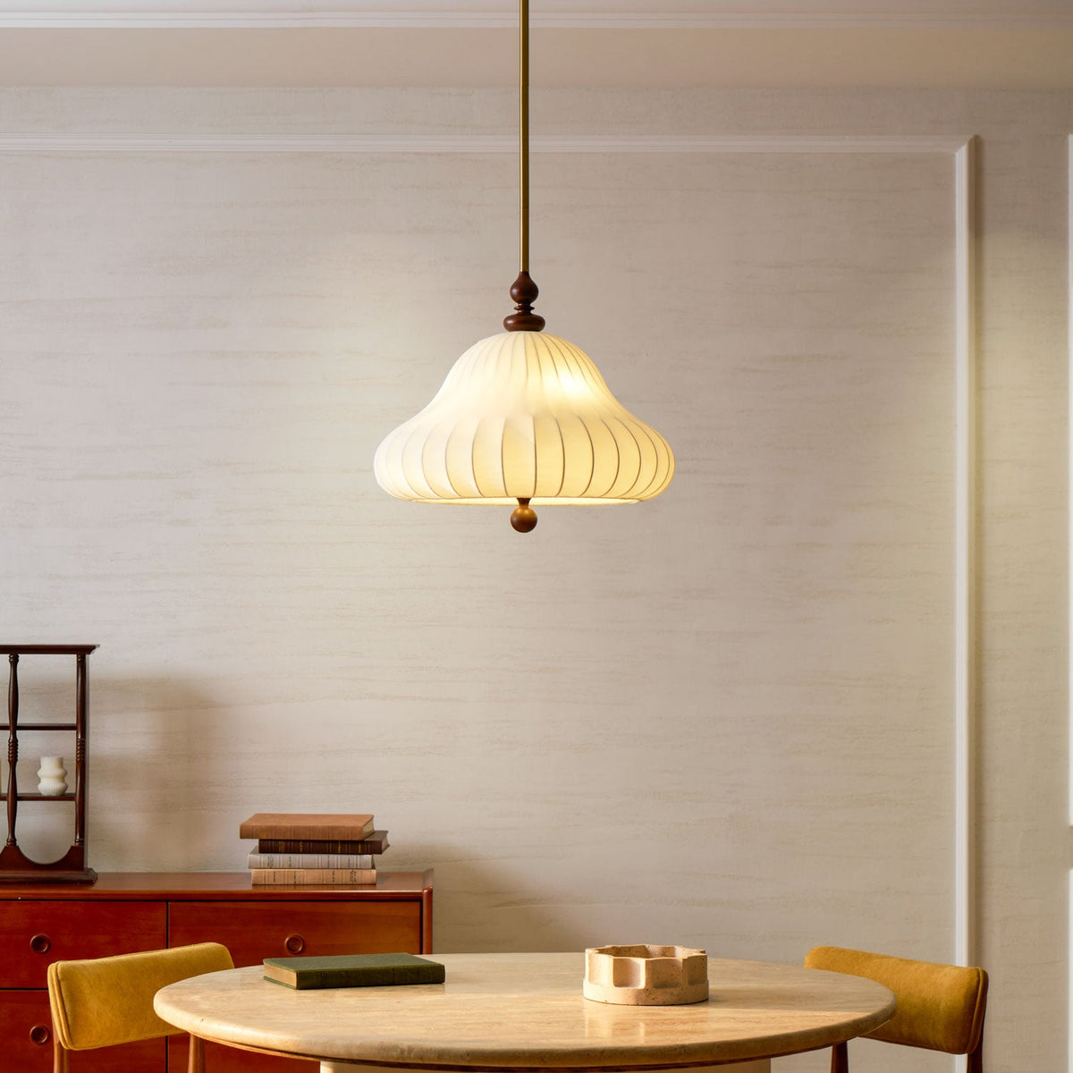 This Clérachic pendant glows warm to the left of a red-brown sideboard, with a round table and yellow chairs below it.