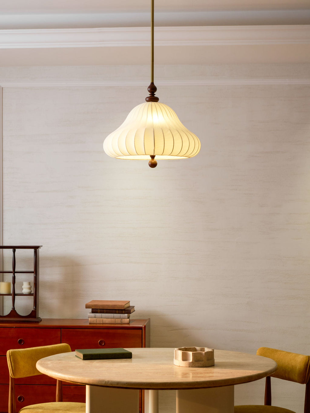 A Clérachic pendant lit above a round travertine-topped table, with yellow chairs and a book stack on the sideboard behind.