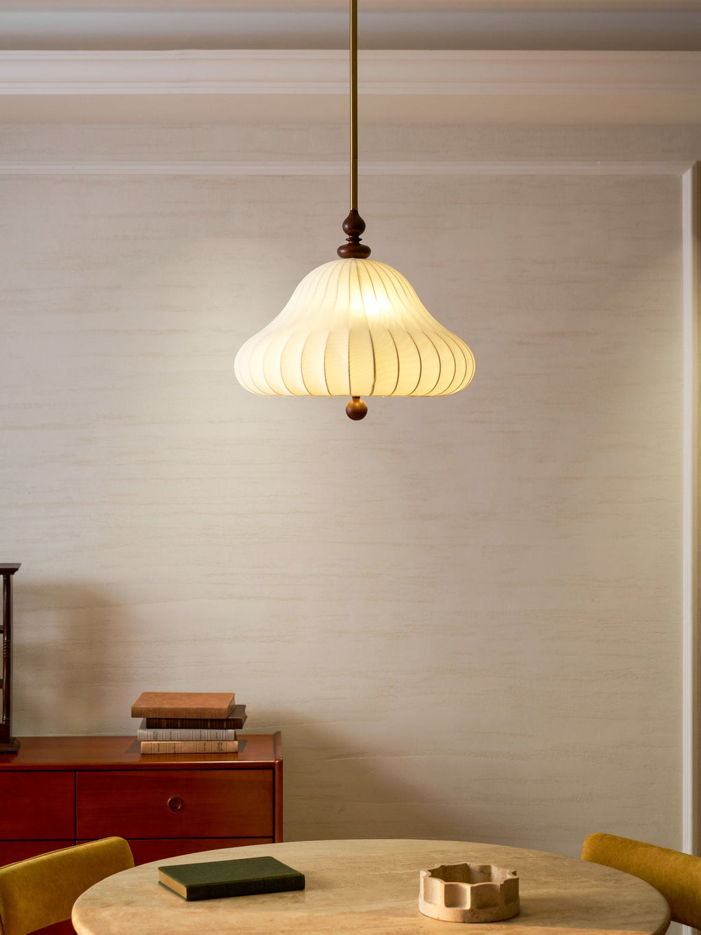 Seen against a pale panelled wall, this Clérachic pendant is lit above a round table set with a green book and a small ceramic bowl.