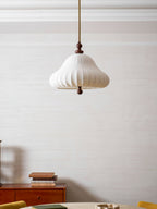 Pleated white shade of this Clérachic pendant seen unlit, hanging on a long rod beside a sideboard with stacked books.