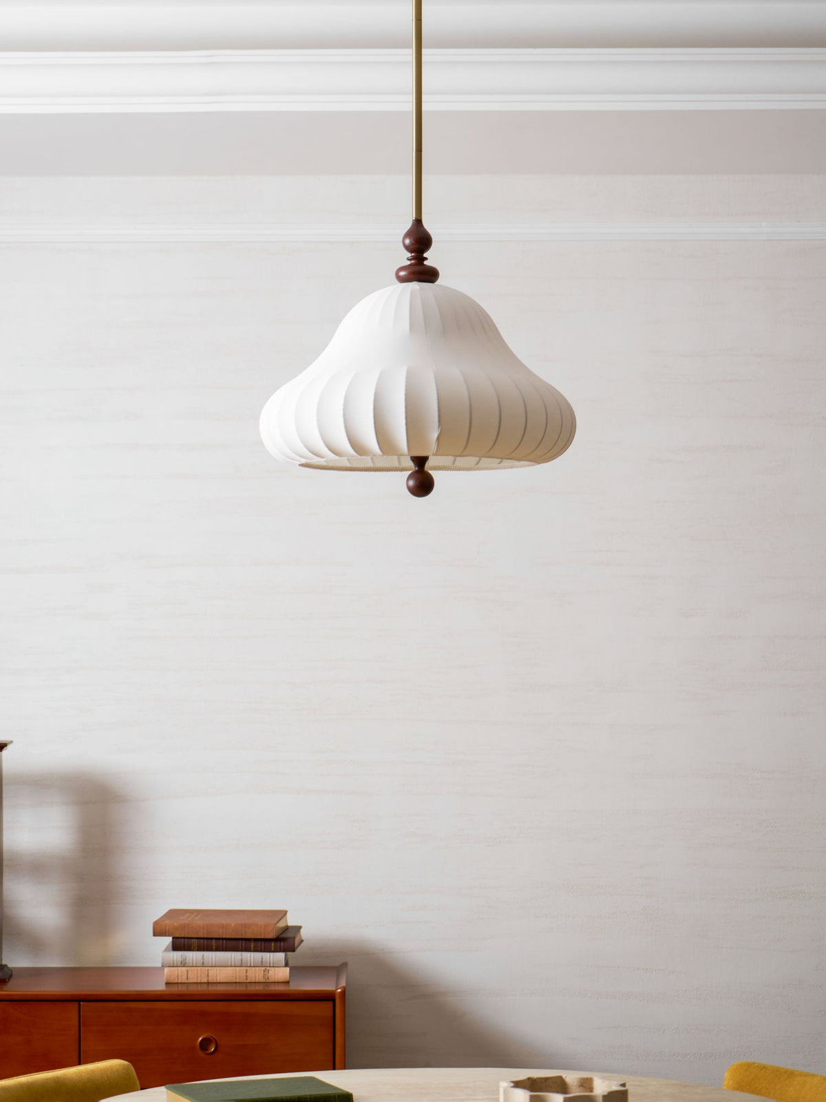 Pleated white shade of this Clérachic pendant seen unlit, hanging on a long rod beside a sideboard with stacked books.