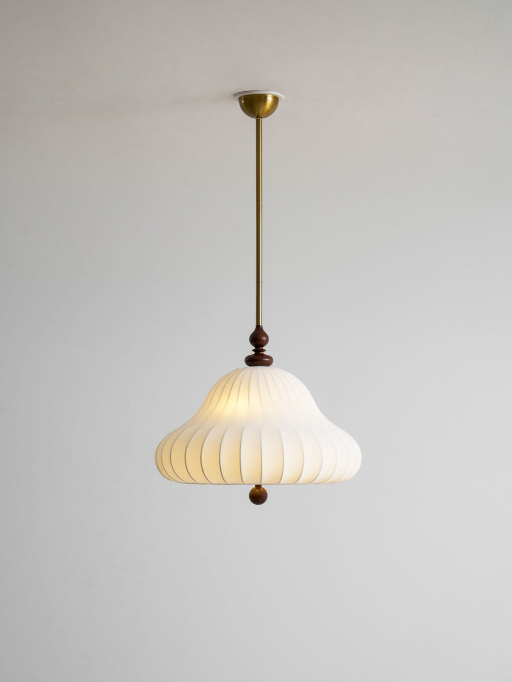 Full length of this Clérachic pendant on a plain grey wall, with a small brass canopy, a long rod and a softly lit white shade.