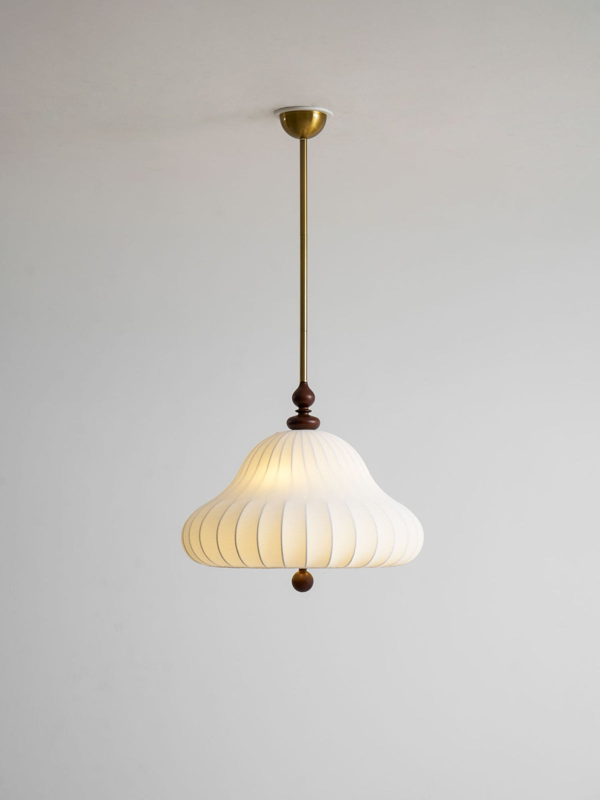 Full length of this Clérachic pendant on a plain grey wall, with a small brass canopy, a long rod and a softly lit white shade.