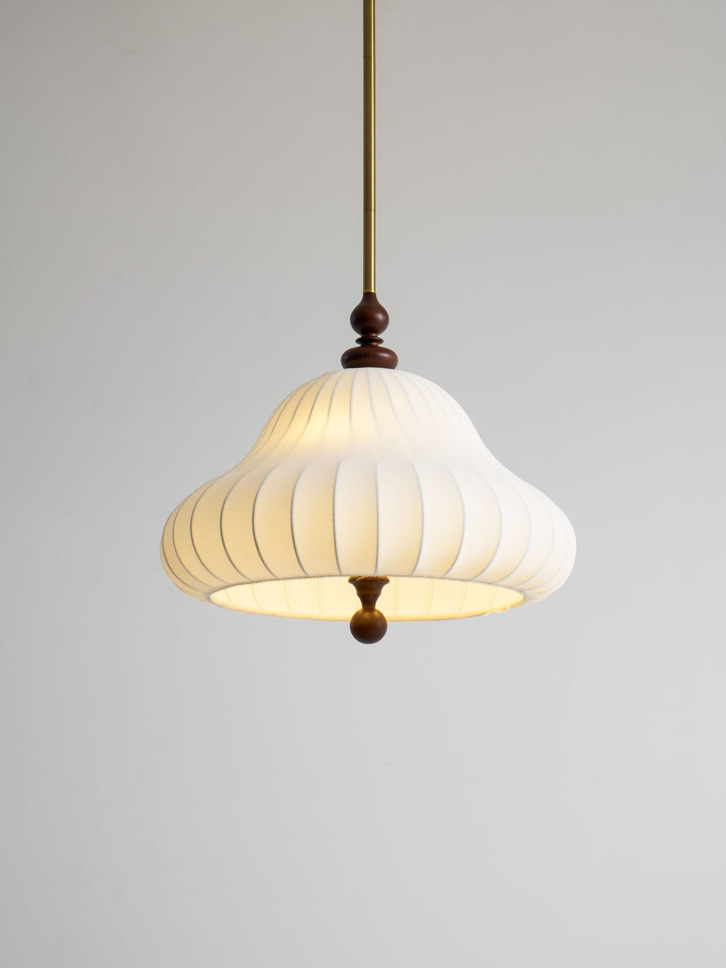 Close angled view of the lit white pleated shade on this Clérachic pendant, with its brass rod and walnut detail on a grey backdrop.